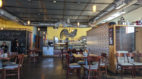 Interior of a cozy restaurant featuring wooden tables, a large rooster mural, and a display case with baked goods.